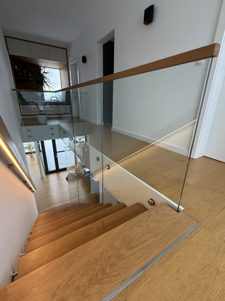 Eine moderne Holztreppe mit Glasgeländer und eingebauter LED-Beleuchtung, die zu einem Flur mit Holzböden, weißen Wänden und minimalistischem Dekor führt.