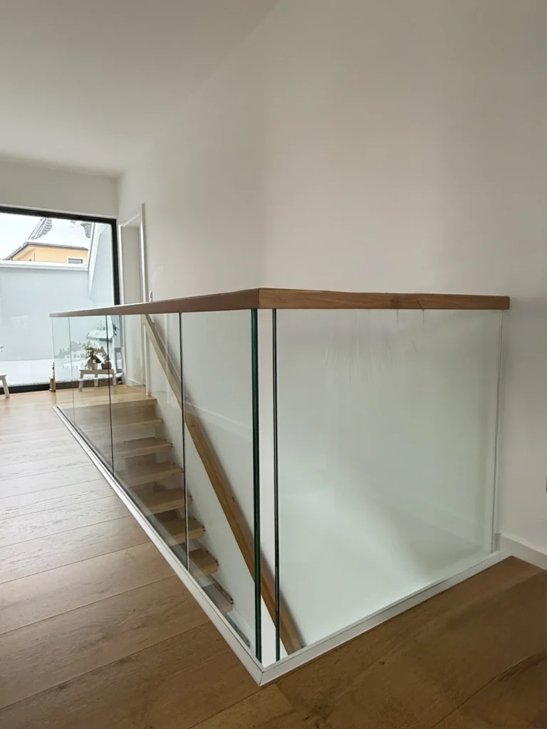 Eine moderne Treppe mit Holzstufen und einem Glasgeländer, gekrönt von einem Holzhandlauf, in einem hellen, minimalistischen Interieur mit weißen Wänden und hellem Holzboden.