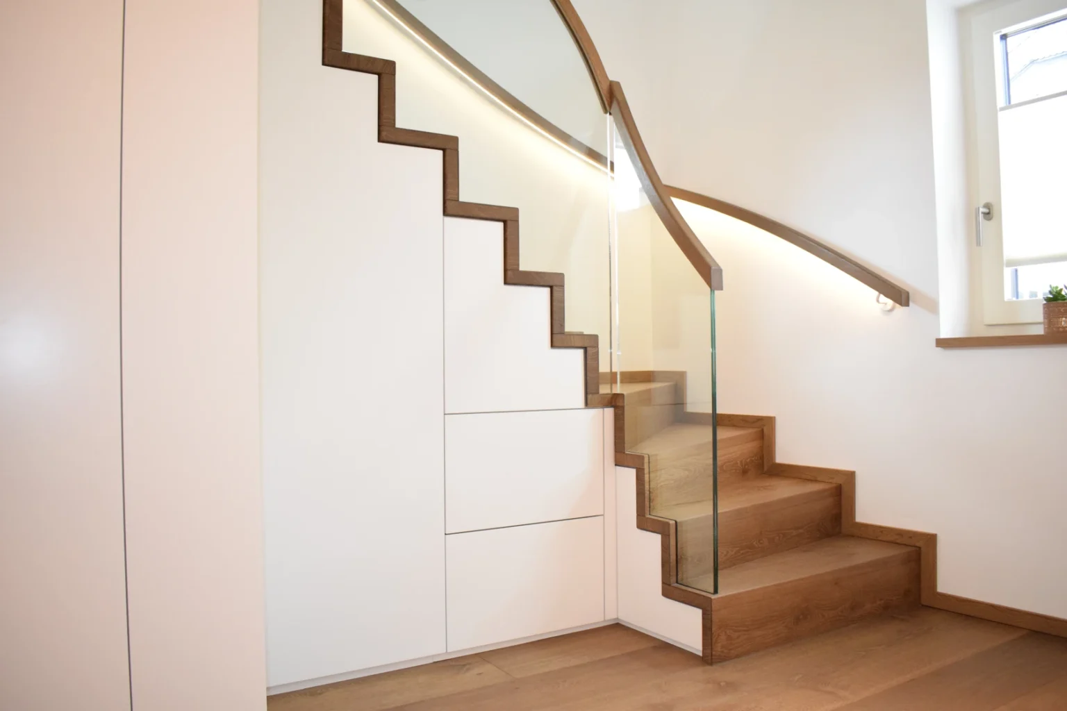 Eine moderne Holztreppe mit eingebauten weißen Schränken darunter, mit einem Glasgeländer und einem Fenster, das Tageslicht hereinlässt.