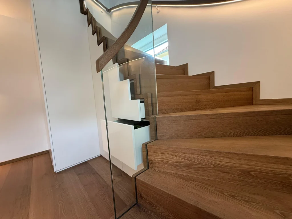 Eine moderne Holztreppe mit Glasgeländer und schlanken Metallstützen, die sich in einem hellen Innenraum mit weißen Wänden und minimalistischem Design nach oben schlängelt.