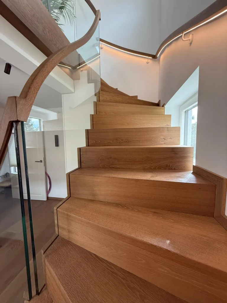 Eine moderne Holztreppe mit scharfen, geometrischen Stufen schlängelt sich entlang eines Glasgeländers und LED-Beleuchtungsbändern in einem hellen Innenraum mit weißen Wänden und großen Fenstern nach oben.