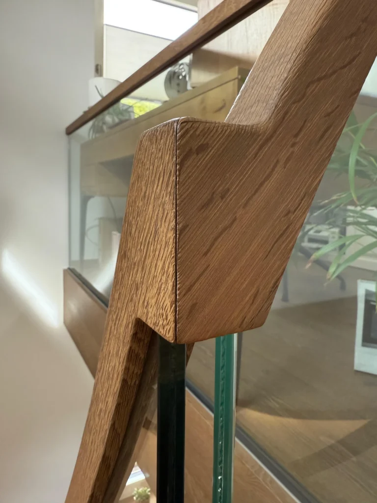 Nahaufnahme eines modernen Holzhandlaufs mit schlankem, kantigem Design, der auf Glasplatten montiert ist. Das Sonnenlicht hebt die Holzmaserung hervor, und im Glas sind Spiegelungen zu sehen. Im Hintergrund sind grüne Pflanzenblätter zu sehen.
