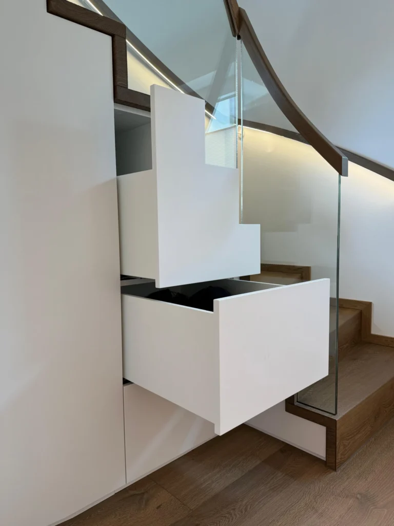 Zwei weiße Schubladen mit unregelmäßigen geometrischen Formen sind in den Raum unter einer Treppe mit Holzstufen und einem Glasgeländer eingebaut. Die Schubladen sind teilweise geöffnet und zeigen eine clevere Nutzung des Stauraums.