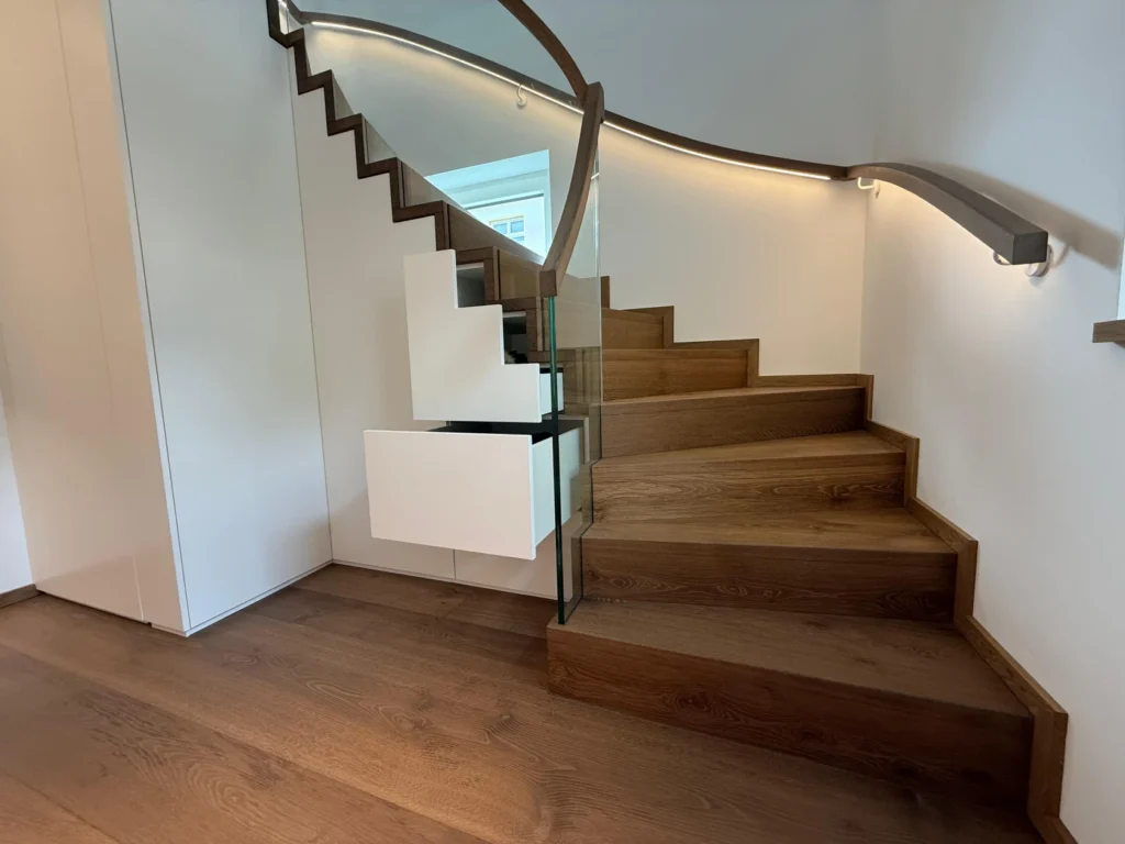 Eine moderne Holztreppe mit Glasgeländer und eingebauten weißen Schubladen darunter, beleuchtet von einer eleganten LED-Handlaufleuchte an der Wand.