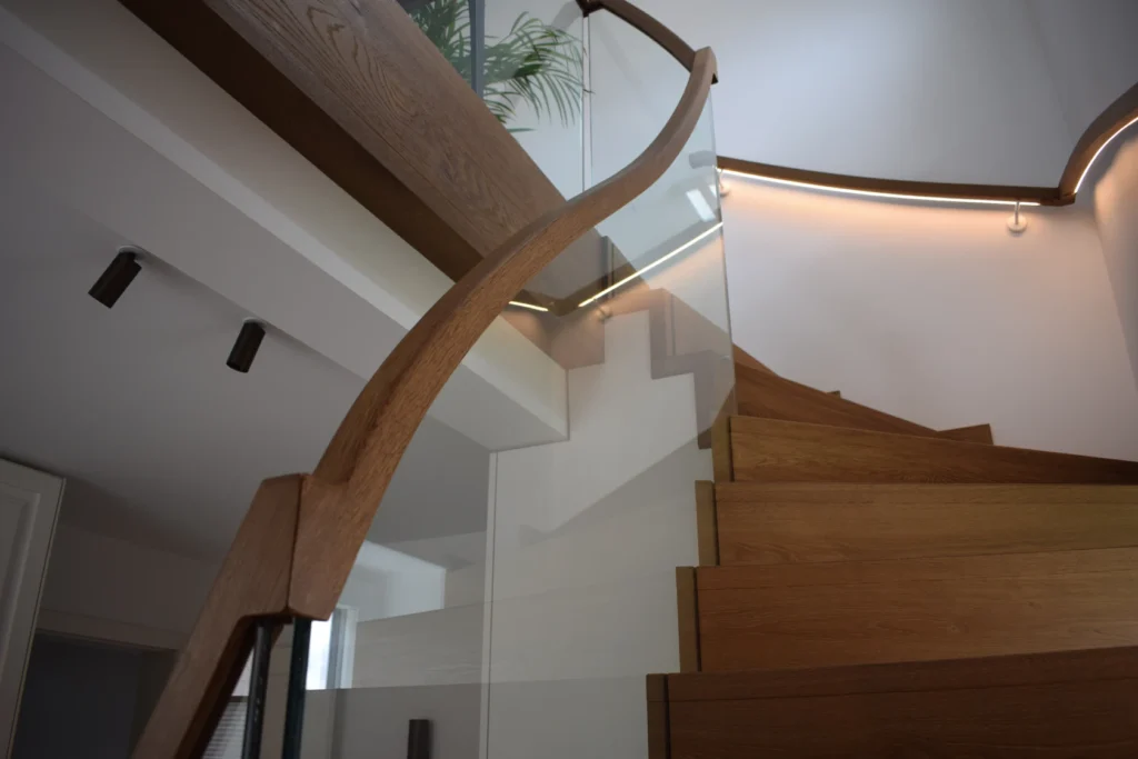 Eine moderne Holztreppe mit geschwungenem Handlauf und Glaspaneelen, die von LED-Lichtbändern entlang der Wand beleuchtet wird. Eine Grünpflanze ist teilweise am oberen Ende der Treppe zu sehen.