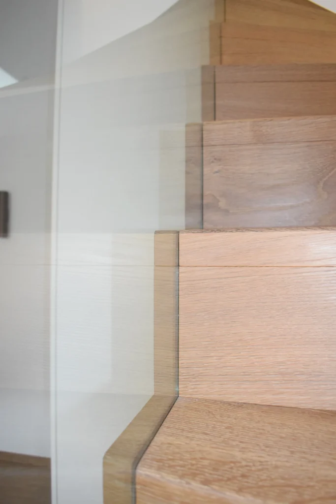 Nahaufnahme einer modernen Holztreppe mit hellbraunen Stufen und einem Glasgeländer vor einer weißen Wand in einem gut beleuchteten Innenraum.