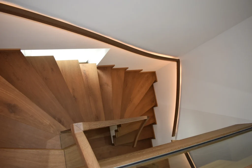 Eine moderne Holzspindeltreppe mit eingebauten LED-Lichtbändern entlang des geschwungenen Handlaufs, von oben betrachtet vor weißen Wänden.