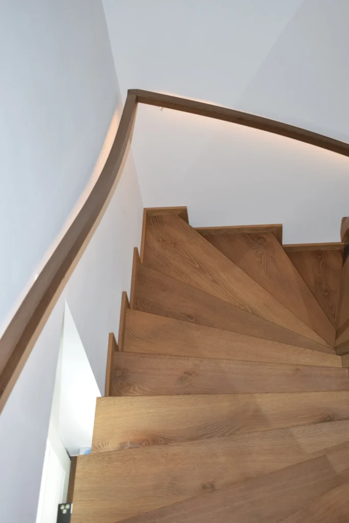Draufsicht auf eine moderne Holzspindeltreppe mit schrägen Stufen, weißen Wänden und einem geschwungenen Holzhandlauf.