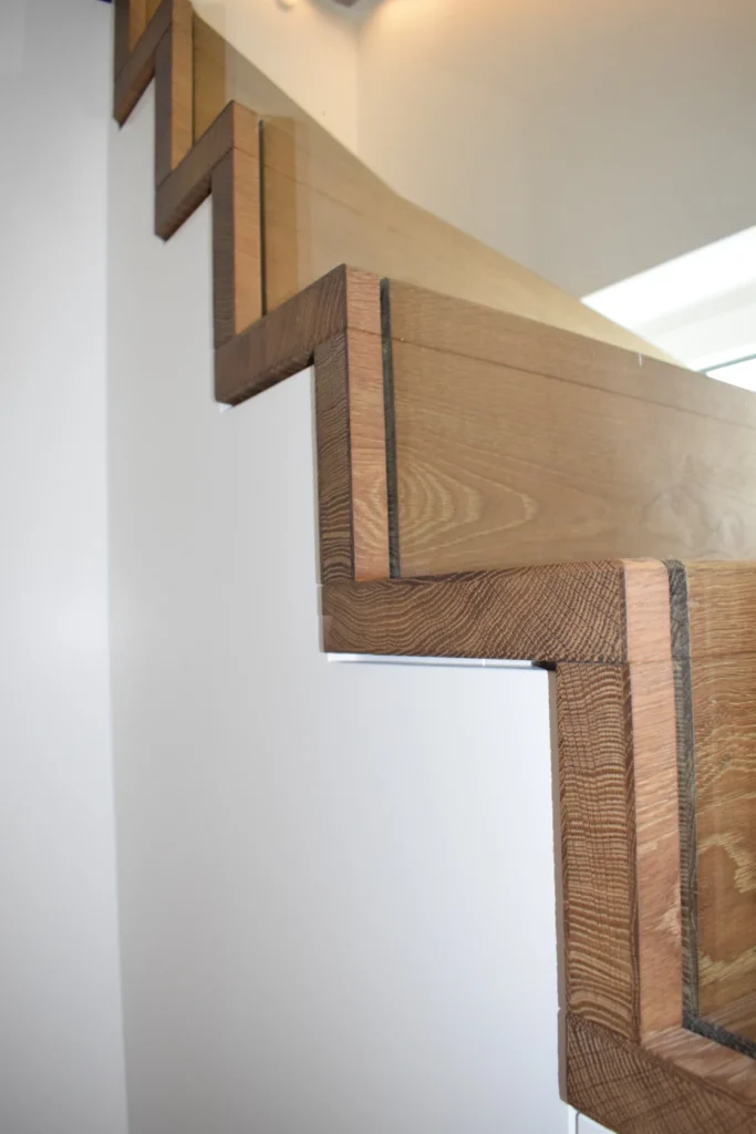 Nahaufnahme einer modernen Holztreppe mit geometrischen, rechteckigen Stufen vor einer weißen Wand. Die Treppe hat klare Linien und ein minimalistisches Design.