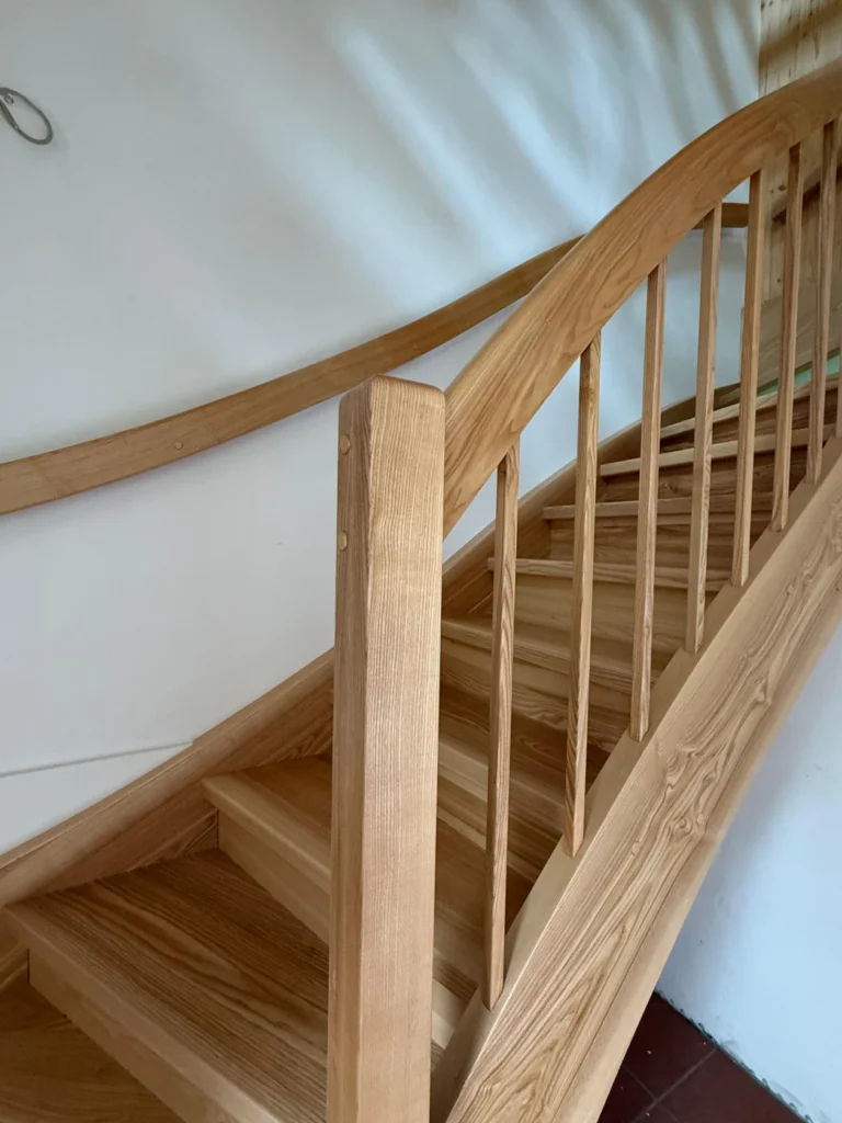 Nahaufnahme einer Holztreppe mit glatten, geschwungenen Handläufen und vertikalen Balustern vor einer weißen Wand. Das Sonnenlicht erzeugt weiche Schatten an der Wand.