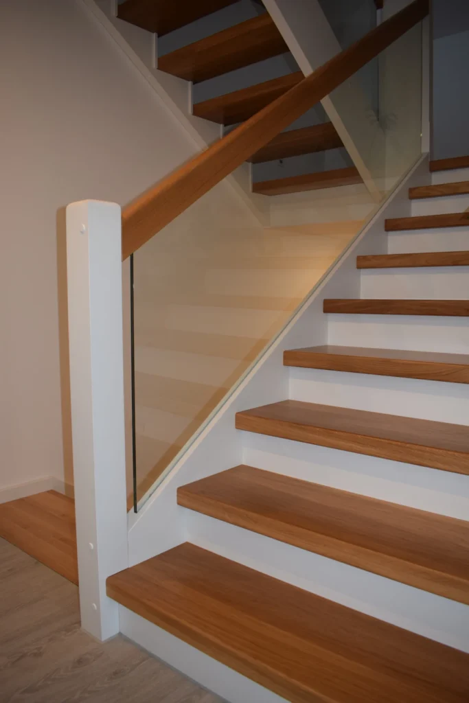 Holztreppe mit weißen Setzstufen und Holzstufen, mit einer Glasplatte und einem Holzhandlauf als modernes Geländer. Die Treppe befindet sich in einem Innenraum und steht vor einer hellen Wand.
