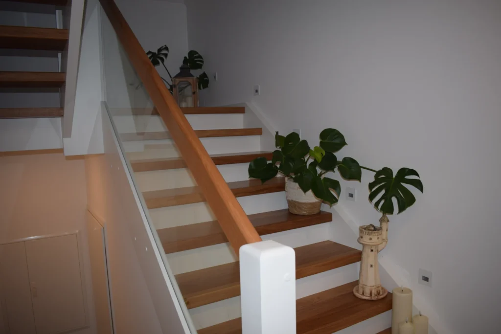 Eine moderne Holztreppe mit Glasgeländer, dekoriert mit grünen Topfpflanzen und einer Holzlaterne, vor weißen Wänden in einem hellen Wohnbereich.