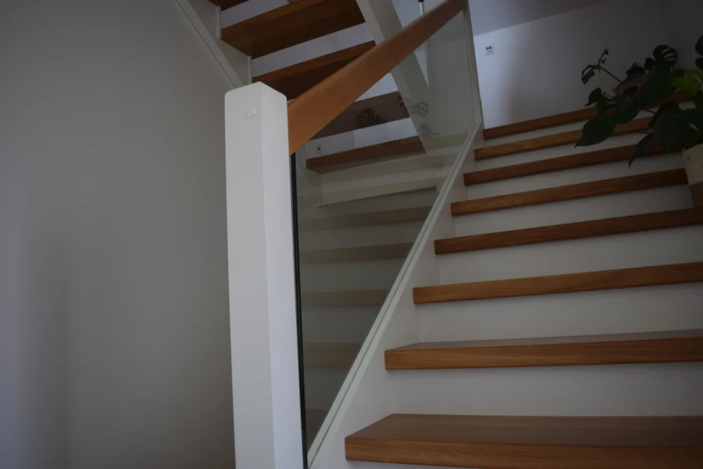 Eine moderne Holztreppe mit weißen Wangen und Glasgeländer führt nach oben. In der oberen rechten Ecke steht eine Topfpflanze, und Licht von oben erhellt den minimalistischen Raum.