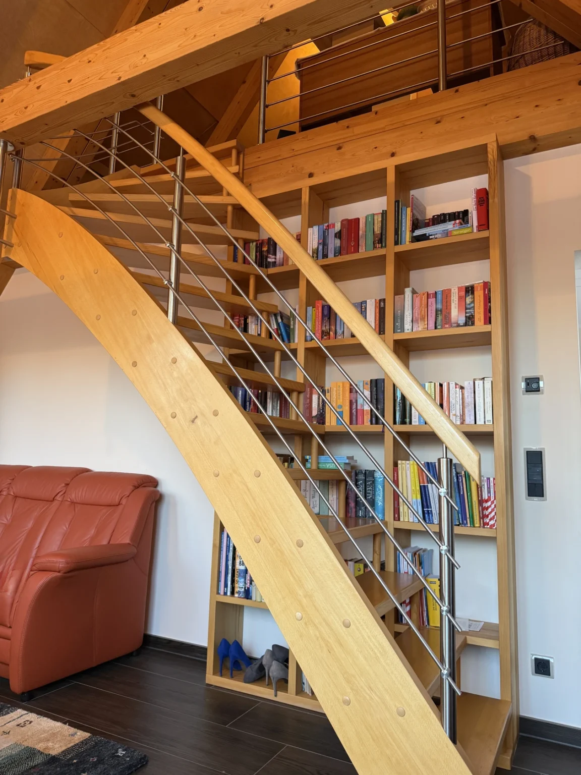 Eine moderne Holztreppe mit Metallgeländer verläuft diagonal über einem eingebauten Bücherregal, das mit Büchern und ein paar Gegenständen gefüllt ist. Unter der Treppe sind ein braunes Ledersofa und ein Paar blaue Schuhe zu sehen.