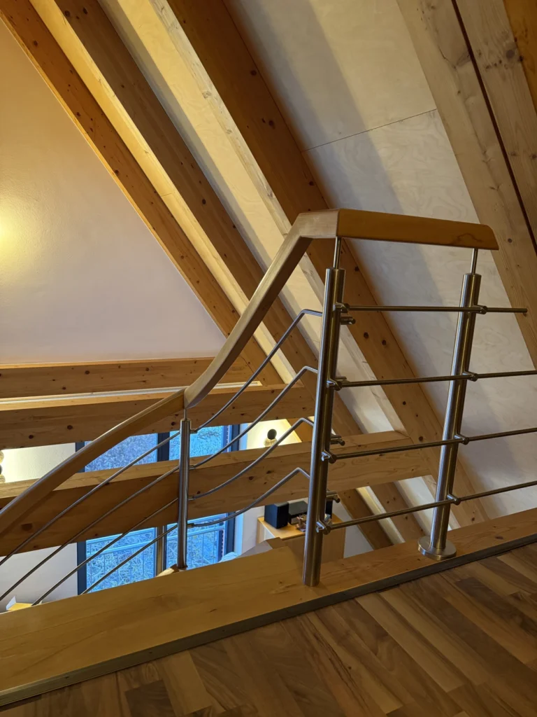 Ein modernes Holztreppengeländer mit Edelstahlseilhalterungen in einem Loft mit freiliegenden Holzbalken und einer schrägen Decke. Im Hintergrund sind eine sanfte gelbe Beleuchtung und ein Fenster zu sehen.