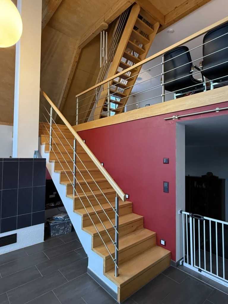 Eine moderne Holztreppe mit Metallgeländer führt zu einem Loftbereich mit zwei schwarzen Stühlen. Die Wand ist rot gestrichen und der Boden ist gefliest. Ein Babygitter befindet sich unten rechts neben einer Tür.