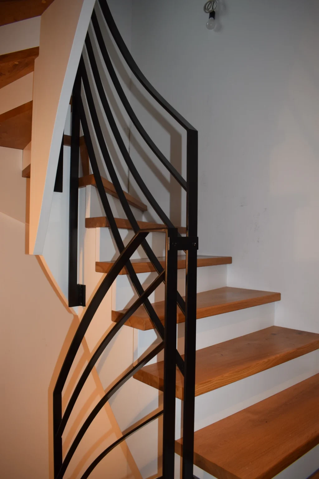 Eine moderne Holztreppe mit offenen Setzstufen und einem schwarzen Metallgeländer mit geometrischen Mustern vor einer weißen Wand.