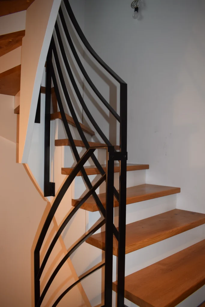Eine moderne Holztreppe mit offenen Setzstufen und einem schwarzen Metallgeländer mit geometrischen Mustern vor einer weißen Wand.