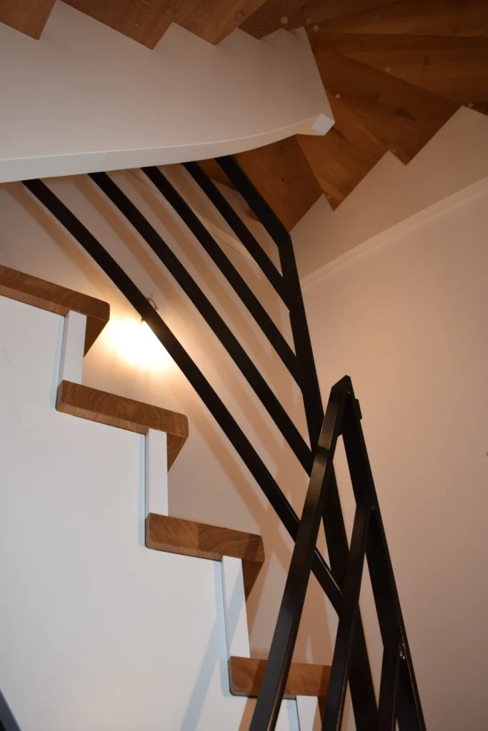 Eine moderne, gewendelte Holztreppe mit schwarzem Metallgeländer schlängelt sich vor einer weißen Wand nach oben; die Stufen haben kontrastierende Holzstufen und weiße Setzstufen.