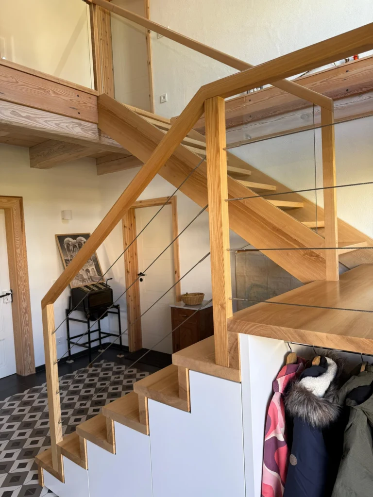 Eine moderne Holztreppe mit Glas- und Seilgeländer, die zu einem oberen Loftbereich führt. Unter der Treppe ist ein Stauraum eingebaut und die Mäntel hängen an Haken in der Nähe. Der Boden ist mit einem geometrischen Fliesenmuster versehen.