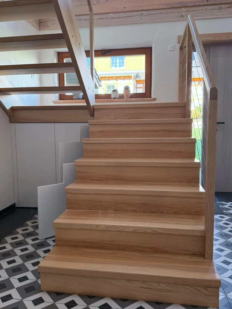 Eine helle Holztreppe mit offenen Setzstufen führt zu einem oberen Stockwerk in einem modernen Haus. Der Boden ist mit geometrischen schwarzen und weißen Fliesen ausgelegt, und das Sonnenlicht fällt durch ein Fenster und eine Glastür im Hintergrund ein.