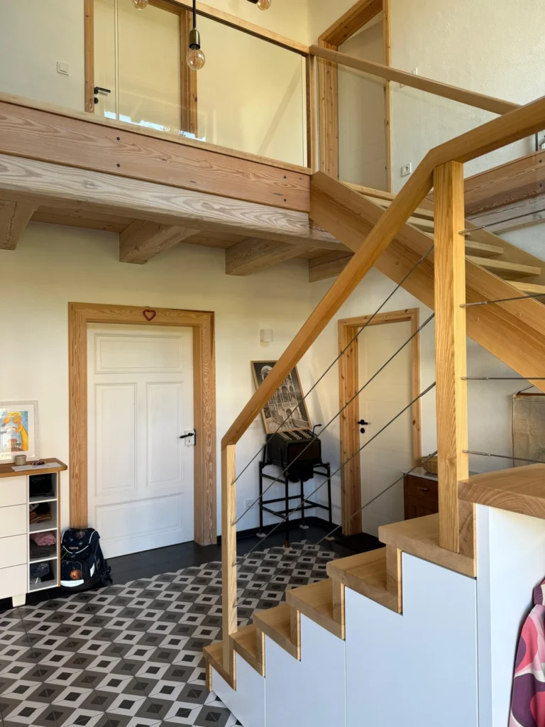 Modernes Interieur mit Holztreppe und Glasgeländer, geometrischem schwarz-weißem Fliesenboden, weißen Türen mit Holzrahmen und minimalistischen Möbeln, darunter ein Schuhregal und ein Beistelltisch.