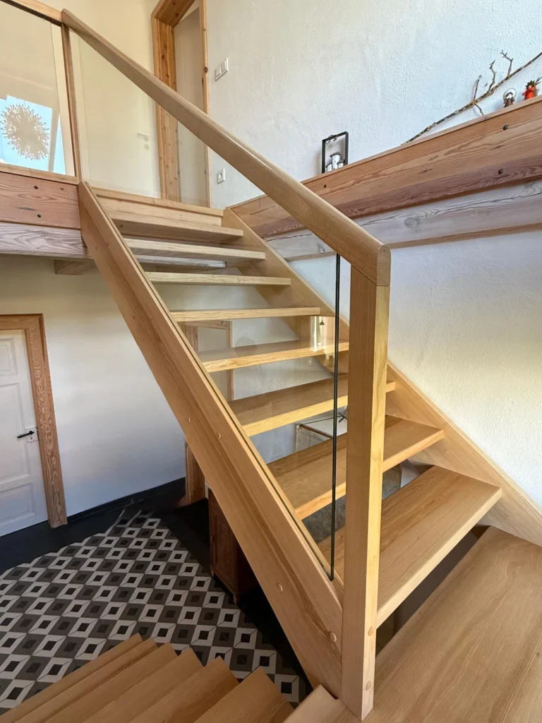 Moderne Holztreppe mit offenen Setzstufen, Glaswangen und Handläufen aus Naturholz in einem hellen Innenraum. Die Treppe führt von einem geometrisch gemusterten Fliesenboden zu einem Flur mit Holztürrahmen.