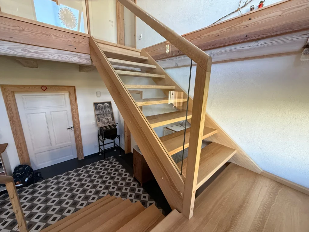 Eine moderne Holztreppe mit Glasgeländer und offenen Setzstufen, die in ein Obergeschoss führt. Darunter befinden sich ein gemusterter Fliesenboden, eine weiße Tür und ein kleiner Holzstuhl an der Wand.