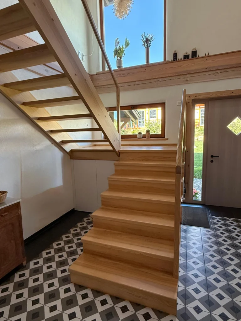 Eine moderne Holztreppe mit einfachem Geländer steht in einem hellen Eingangsbereich mit gemusterten schwarz-weißen Bodenfliesen, großen Fenstern und Topfpflanzen auf einem Sims darüber.