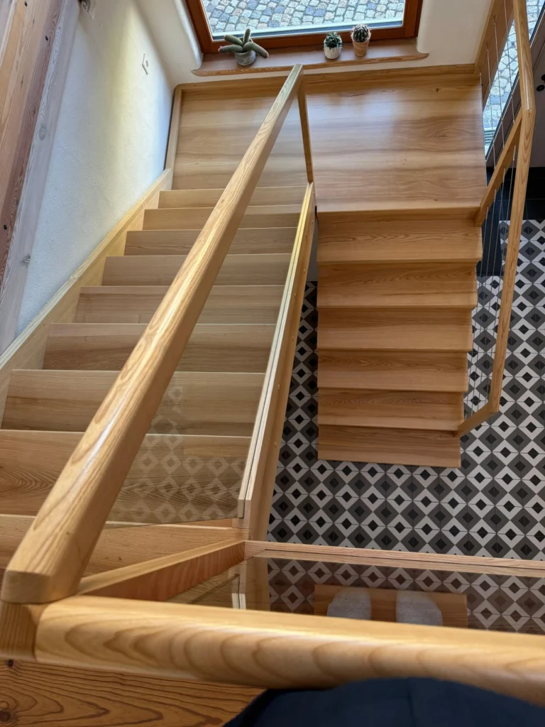 Blick auf eine moderne Holztreppe mit Glasgeländer, die zu einem schwarz-weiß geometrisch gemusterten Fliesenboden führt, neben einem Fenster mit drei kleinen Topfpflanzen auf der Fensterbank.