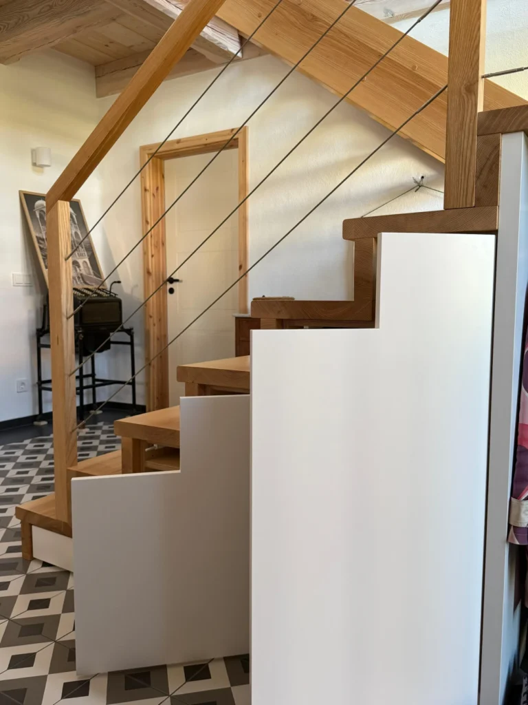 Eine moderne Holztreppe mit Seilgeländer, geometrischen weißen Seitenwänden und gemustertem schwarz-weißem Fliesenboden in einem hellen Innenraum.