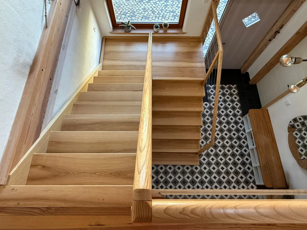 Blick von oben auf eine Holztreppe, die zu einem gefliesten Eingangsbereich mit einem geometrischen Schwarz-Weiß-Muster, einem großen Fenster und Holzbalken an den Wänden hinunterführt.