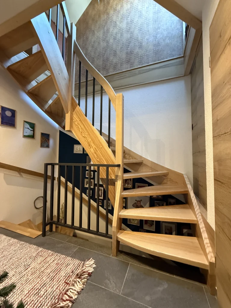 Eine moderne Holztreppe mit schwarzem Metallgeländer in einem gut beleuchteten Innenraum. An den Wänden hängen kleine gerahmte Kunstwerke, und auf dem oberen Treppenabsatz befindet sich eine gemusterte Wand. Die Treppe führt nach oben und mündet in einen Treppenabsatz.