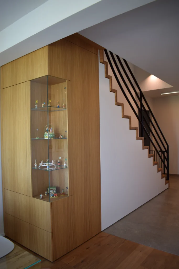 Eine moderne Treppe mit schwarzem Geländer befindet sich über einer eingebauten Holzvitrine mit Glasböden, in der verschiedene kleine Figuren und Sammlerstücke ausgestellt sind. Der Bereich ist gut beleuchtet und minimalistisch gestaltet.