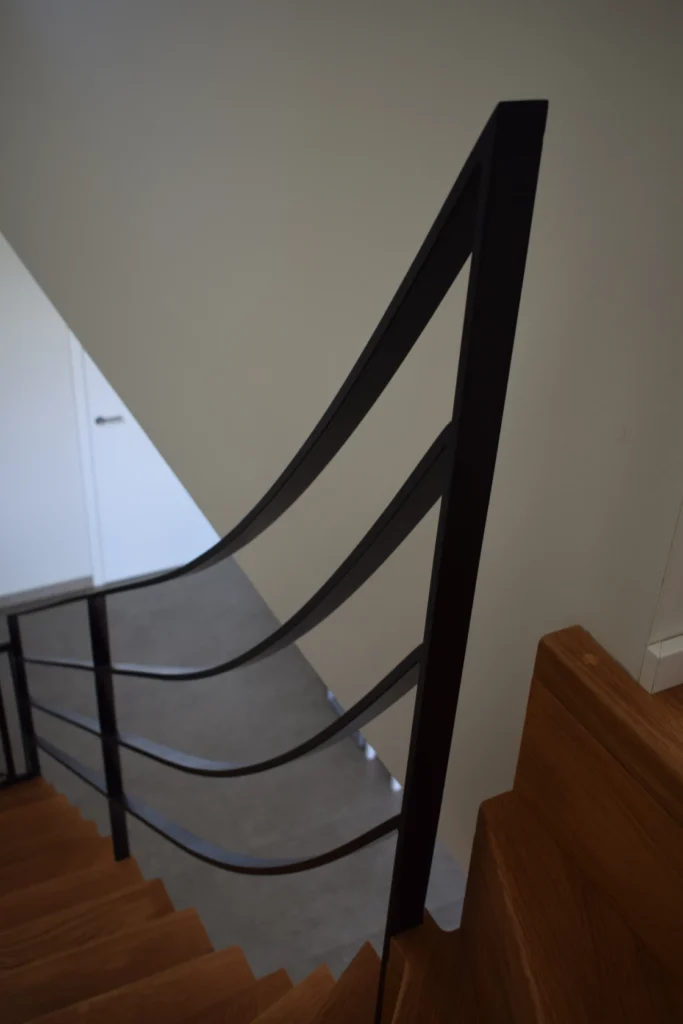 Schwarzes Metallgeländer mit drei geschwungenen Stäben entlang einer Holztreppe, die zu einem grauen Boden und einer weißen Tür in einem modernen, minimalistischen Innenraum hinunterführt.