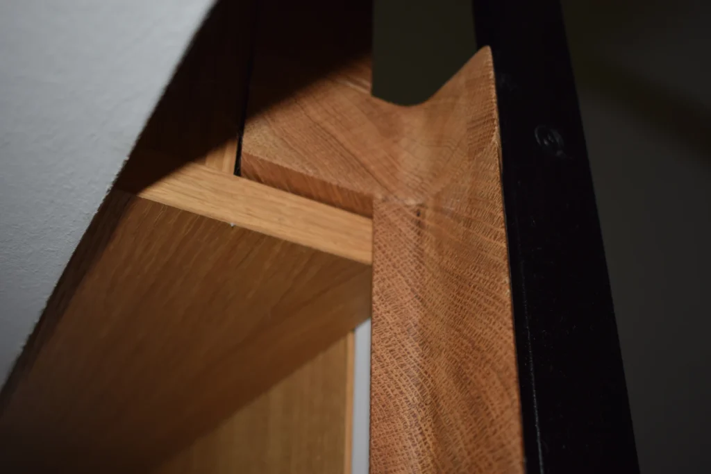 Nahaufnahme der ineinandergreifenden Holzverbindungen eines Möbelstücks, die eine präzise Handwerkskunst mit hellen und dunklen Holztönen und einer schwarzen Metallkante auf der rechten Seite zeigen.