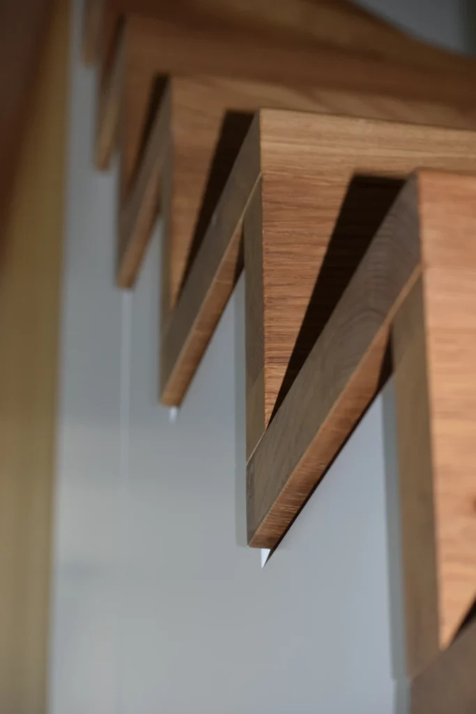 Nahaufnahme der Unterseite einer modernen Holztreppe mit kantigen, zickzackförmigen Stufen vor einer hellgrauen Wand. Die Perspektive unterstreicht das geometrische Design und die klaren Linien der Struktur.