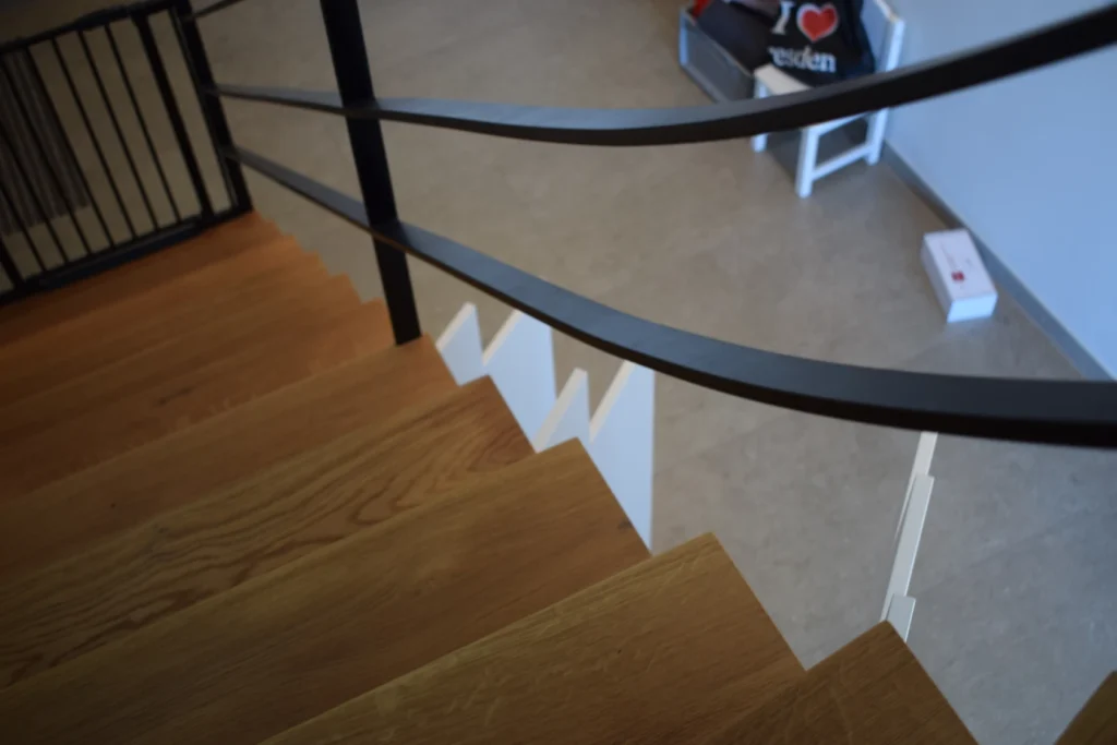 Blick von oben auf eine Holztreppe mit schwarzem Metallgeländer. Unten sind ein beiger Boden, eine schwarze Tasche mit der Aufschrift I ❤️ und ein weißer Kasten zu sehen.