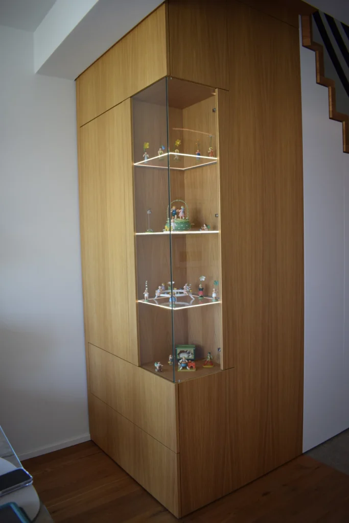 Eine hölzerne Vitrine mit Glastüren, die in einer Zimmerecke eingebaut ist und mehrere kleine Figuren und Modelle enthält, die auf drei beleuchteten Regalen angeordnet sind. Auf der rechten Seite ist teilweise eine Treppe sichtbar.