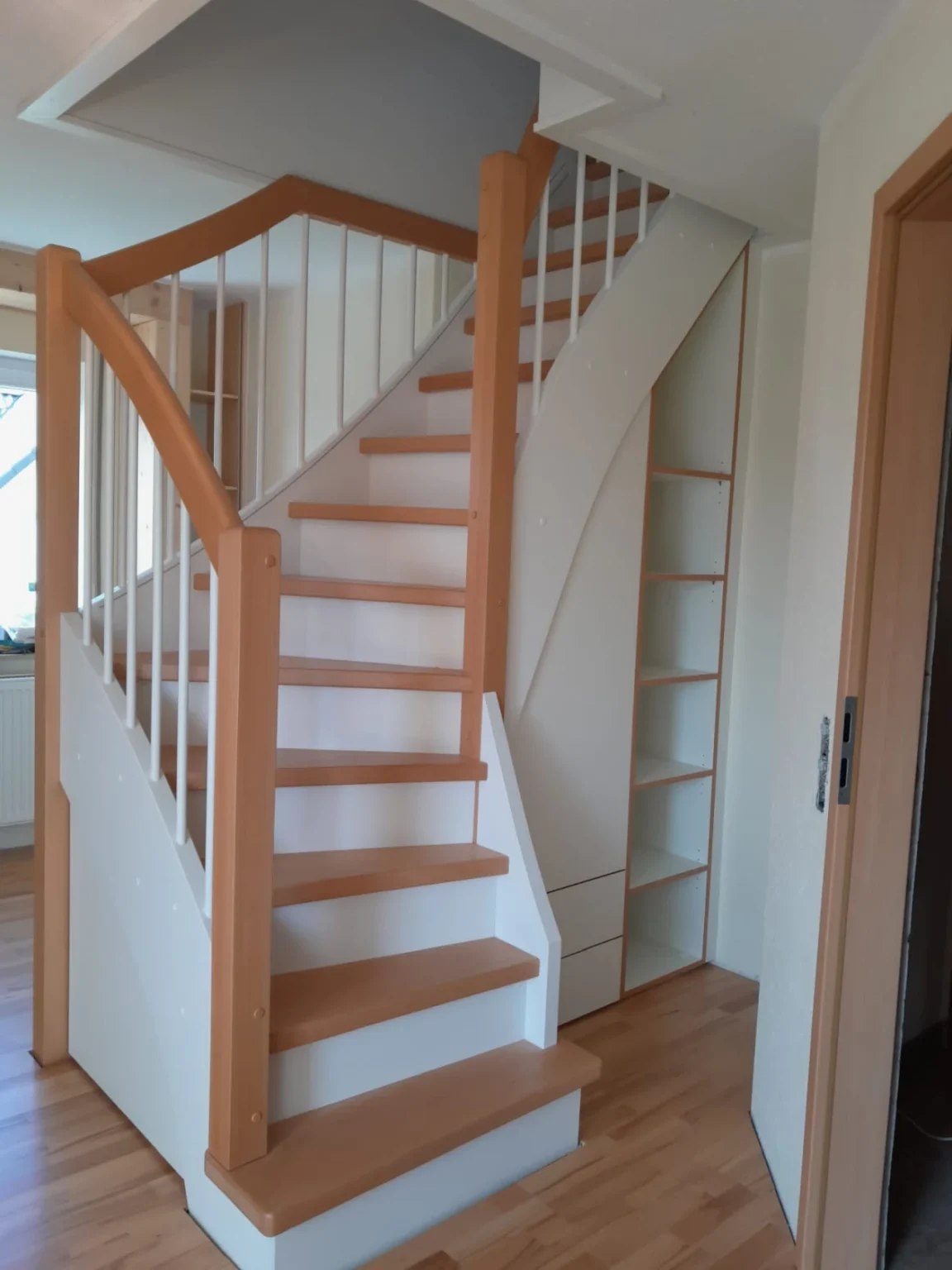 Eine Holztreppe mit hellbraunen Stufen und Handläufen, weißen Geländerstäben und darunter integrierten Ablagefächern in einem Raum mit hellen Wänden und Holzfußboden.