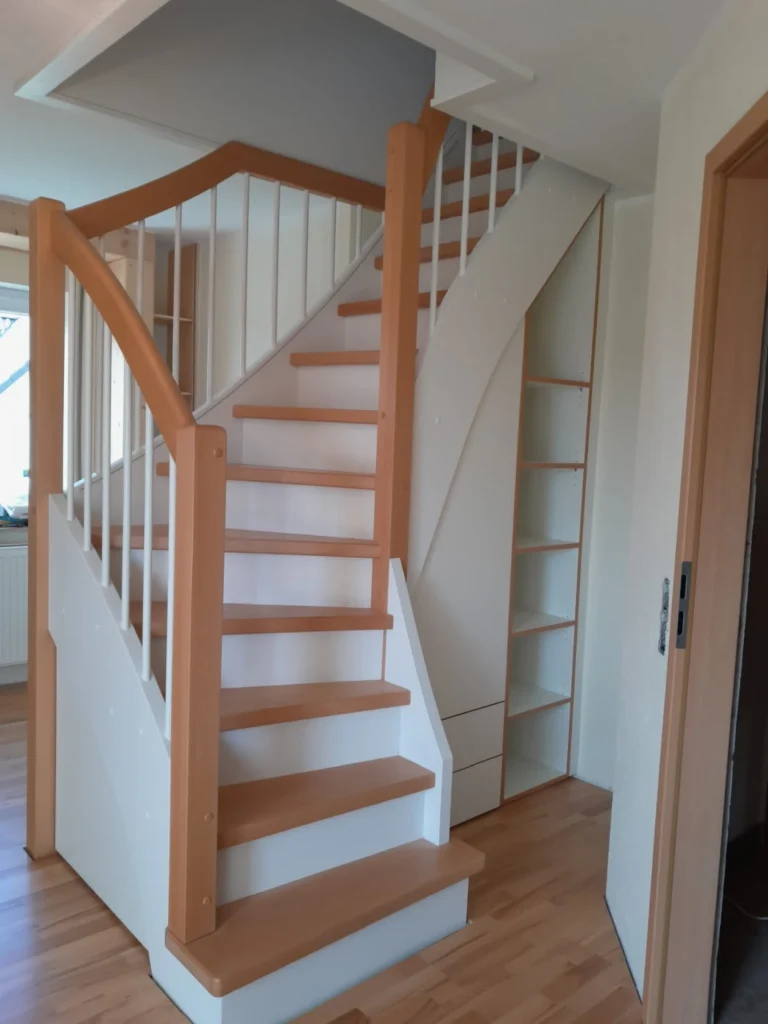 Eine Holztreppe mit hellbraunen Stufen und Handläufen, weißen Geländerstäben und darunter integrierten Ablagefächern in einem Raum mit hellen Wänden und Holzfußboden.
