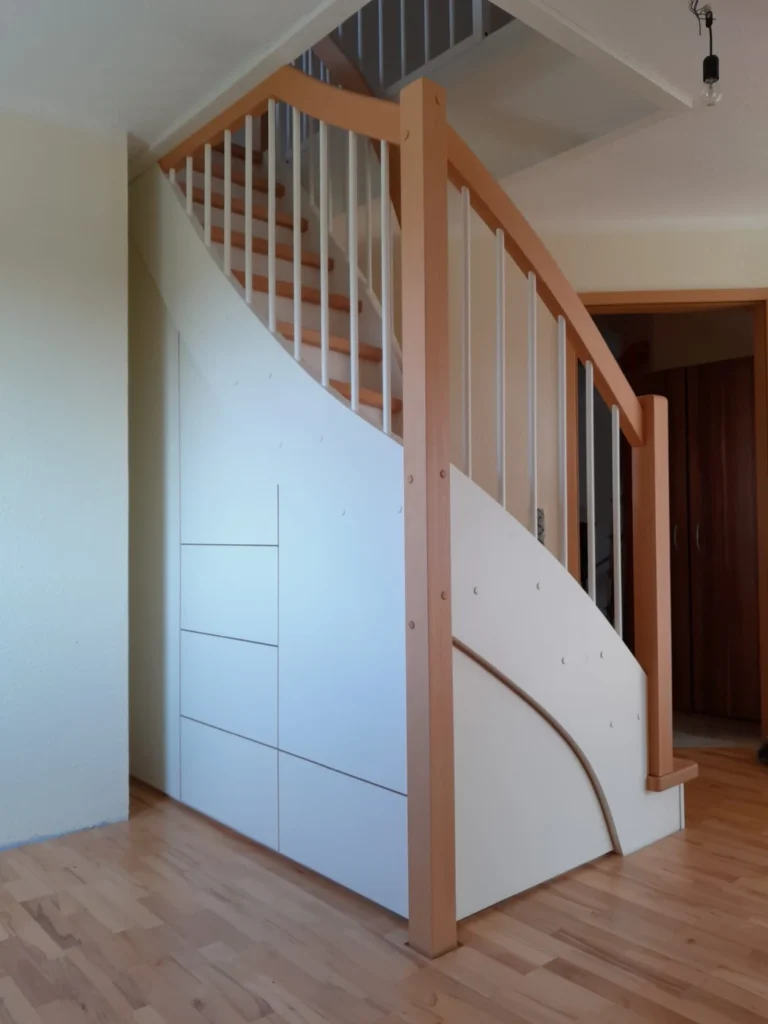 Eine Holztreppe mit weißen Einbauschränken darunter, mit Handläufen aus Naturholz und hellen Wänden und Böden.
