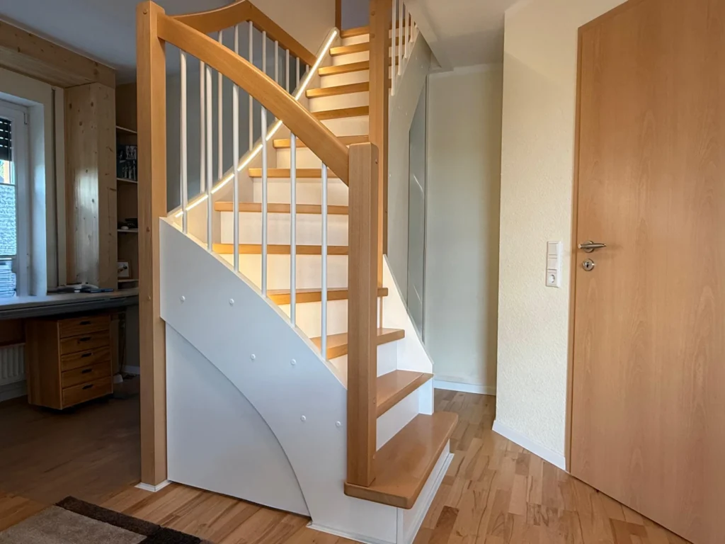 Eine moderne Holztreppe mit weißem Geländer schlängelt sich in einem gut beleuchteten, aufgeräumten Raum mit hellem Holzboden nach oben. Im Hintergrund sind eine geschlossene Holztür und ein Schreibtisch mit Regalen und einem Fenster zu sehen.