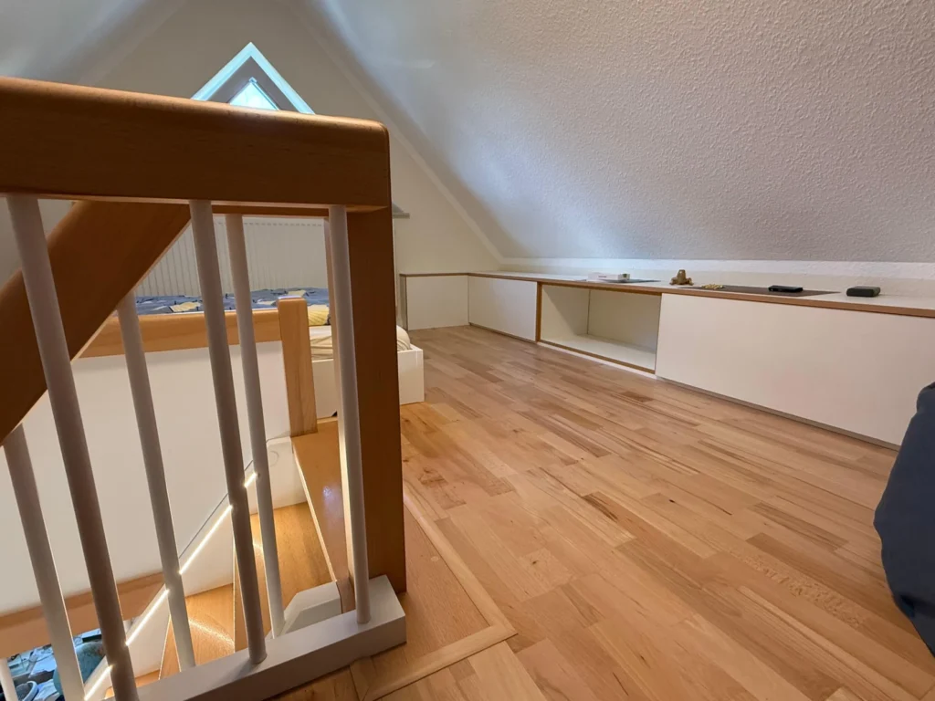 Ein helles Loft mit schrägen Decken, Holzfußböden, weißen Einbauschränken an der Wand und einem Holztreppengeländer im Vordergrund. Ein dreieckiges Fenster lässt natürliches Licht herein.