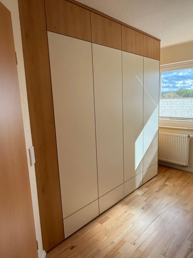 Ein moderner Einbauschrank mit hellen Holz- und weißen Paneeltüren steht an einer Wand in einem hellen Raum mit Holzböden und einem Fenster, das Tageslicht hereinlässt.