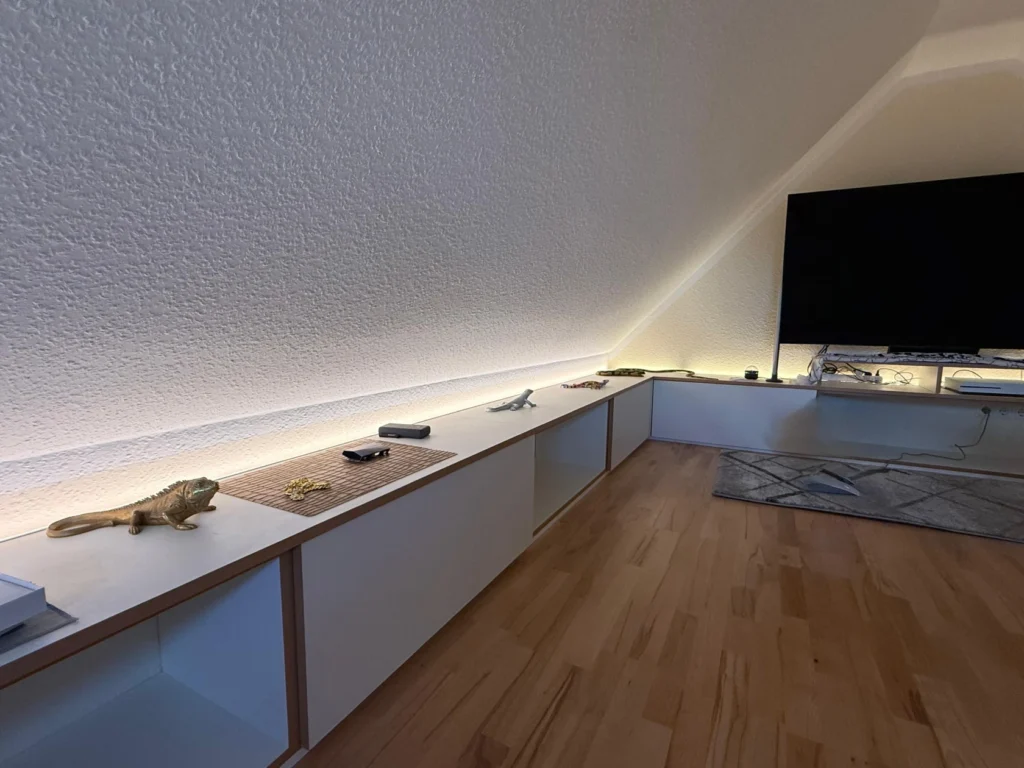 Eine minimalistische Wohnzimmerecke mit weißen, niedrigen Schränken an der Wand, einem Flachbildfernseher, Fernbedienungen und einer Bartagame, die auf dem Schrank sitzt. Hinter den Schränken sind warme LED-Leuchten angebracht. Holzboden und neutrales Dekor.