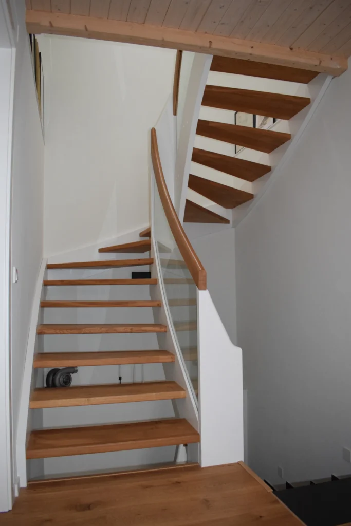 Eine moderne Holztreppe mit offenen Setzstufen und weißen Seitenwangen schlängelt sich in einem hellen, minimalistischen Raum mit weißen Wänden und einer hellen Holzdecke nach oben.