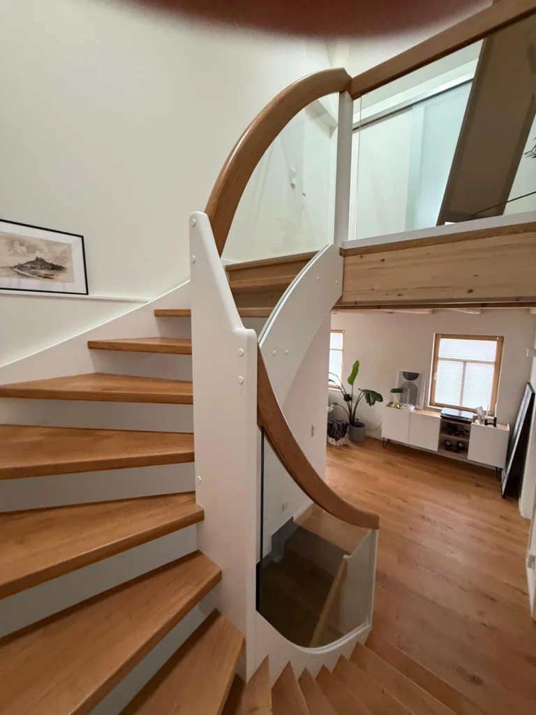 Eine moderne Holztreppe mit weißen Stützen und Glaspaneelen führt zu einem Loft in einem hellen, offenen Wohnbereich mit Holzböden, großen Fenstern und minimalistischem Dekor.
