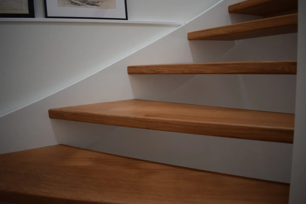 Eine Nahaufnahme einer modernen Holztreppe mit offenen Setzstufen vor einer weißen Wand; gerahmte Bilder sind teilweise an der Wand darüber zu sehen.