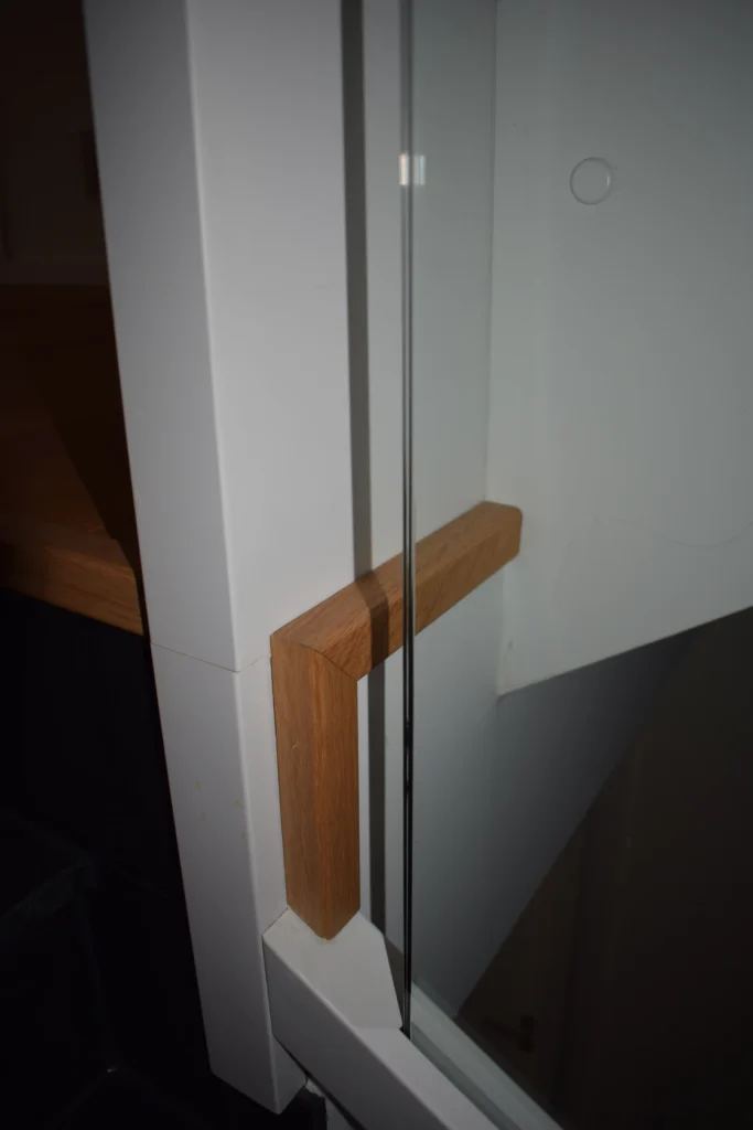 Nahaufnahme einer modernen Treppenecke mit einem Holzhandlauf, der mit einer Glasplatte und einer weißen Wand verbunden ist. Die Materialien unterstreichen ein minimalistisches, zeitgenössisches Design.